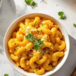 Overhead flat lay of creamy Instant Pot Mac and Cheese on a white ceramic plate with golden cheese sauce, garnished with fresh parsley and shredded cheddar, small bowl of extra cheese sauce on the side, white linen napkin, bright natural daylight on white table