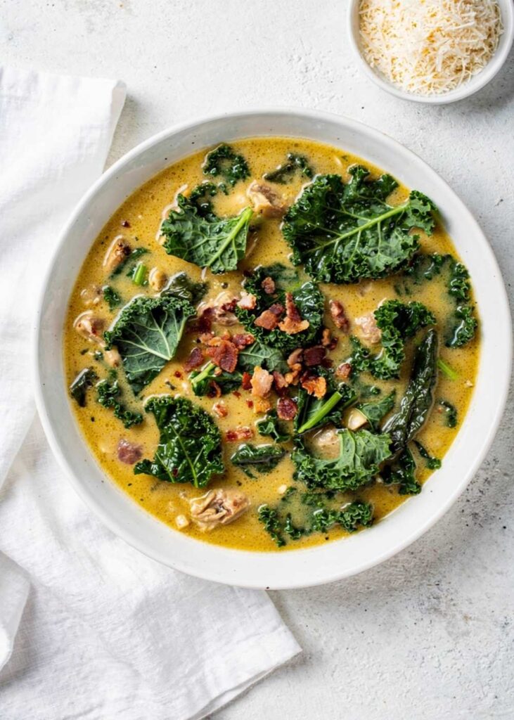 Zuppa Toscana Zuppa Toscana recipe served in a white ceramic bowl overhead flat lay, creamy golden soup with Italian sausage, kale, and crumbled bacon, Parmesan cheese on the side, white marble table, natural daylight