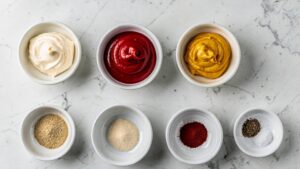 Flat lay overhead shot of all Classic Burger Sauce ingredients arranged in small white bowls on a clean white marble table in bright natural light.