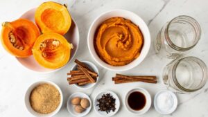 Flat lay overhead shot of all Pumpkin Butter ingredients arranged in white bowls on a clean white marble table in bright natural daylight, including fresh pumpkin, puree, brown sugar, cinnamon, nutmeg, cloves, vanilla, salt, and Mason jars