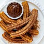 Golden homemade churros arranged on a white ceramic plate dusted with cinnamon sugar, with a small bowl of chocolate dipping sauce on a white table.