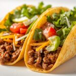 Close-up cross section of a beef taco showing seasoned ground beef filling, shredded cheese, crisp lettuce, and diced tomatoes inside a crispy tortilla shell