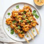 Overhead flat lay of golden bang bang shrimp skewers on a white plate with Bang Bang sauce and cilantro garnish