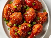 Crispy Gochujang Chicken on a white ceramic plate with glossy red glaze, sesame seeds, green onions, and a small bowl of sauce on a white table.