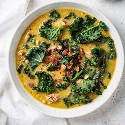 Overhead flat lay of creamy Zuppa Toscana soup in a white ceramic bowl, garnished with fresh kale and crumbled bacon, small bowl of Parmesan cheese on the side, white linen napkin, natural daylight food photography
