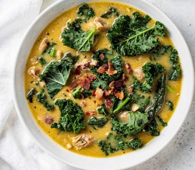Overhead flat lay of creamy Zuppa Toscana soup in a white ceramic bowl, garnished with fresh kale and crumbled bacon, small bowl of Parmesan cheese on the side, white linen napkin, natural daylight food photography