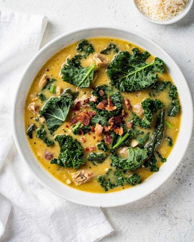 Zuppa Toscana recipe served in a white ceramic bowl overhead flat lay, creamy golden soup with Italian sausage, kale, and crumbled bacon, Parmesan cheese on the side, white marble table, natural daylight