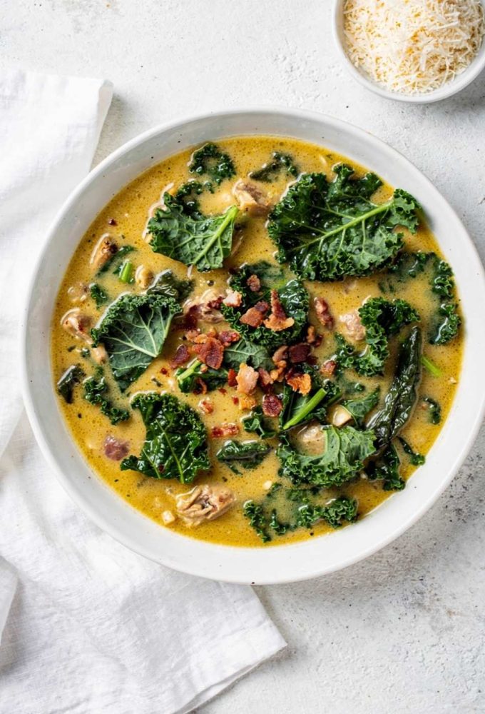Overhead flat lay of creamy Zuppa Toscana soup in a white ceramic bowl, garnished with fresh kale and crumbled bacon, small bowl of Parmesan cheese on the side, white linen napkin, natural daylight food photography