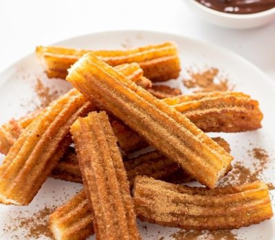Golden homemade churros arranged on a white ceramic plate dusted with cinnamon sugar, with a small bowl of chocolate dipping sauce on a white table.