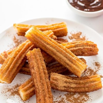 Golden homemade churros arranged on a white ceramic plate dusted with cinnamon sugar, with a small bowl of chocolate dipping sauce on a white table.