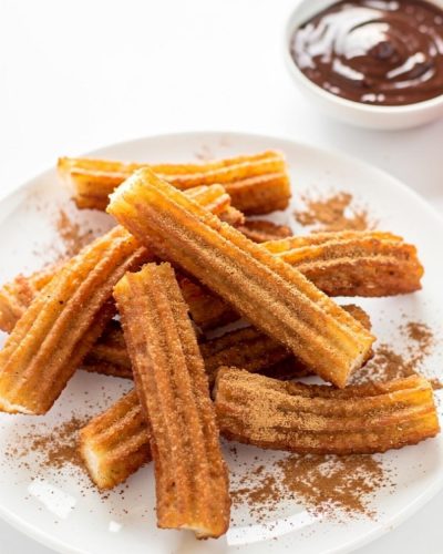 Golden homemade churros arranged on a white ceramic plate dusted with cinnamon sugar, with a small bowl of chocolate dipping sauce on a white table.