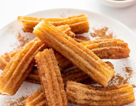 Golden homemade churros arranged on a white ceramic plate dusted with cinnamon sugar, with a small bowl of chocolate dipping sauce on a white table.