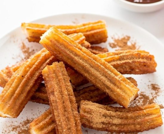 Golden homemade churros arranged on a white ceramic plate dusted with cinnamon sugar, with a small bowl of chocolate dipping sauce on a white table.