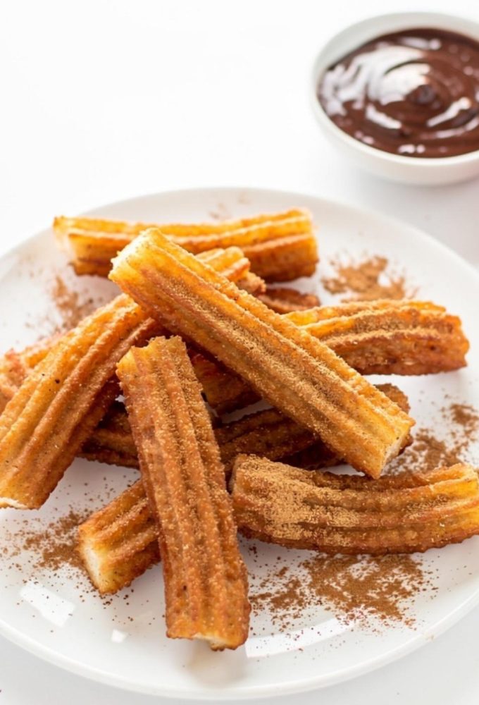 Golden homemade churros arranged on a white ceramic plate dusted with cinnamon sugar, with a small bowl of chocolate dipping sauce on a white table.