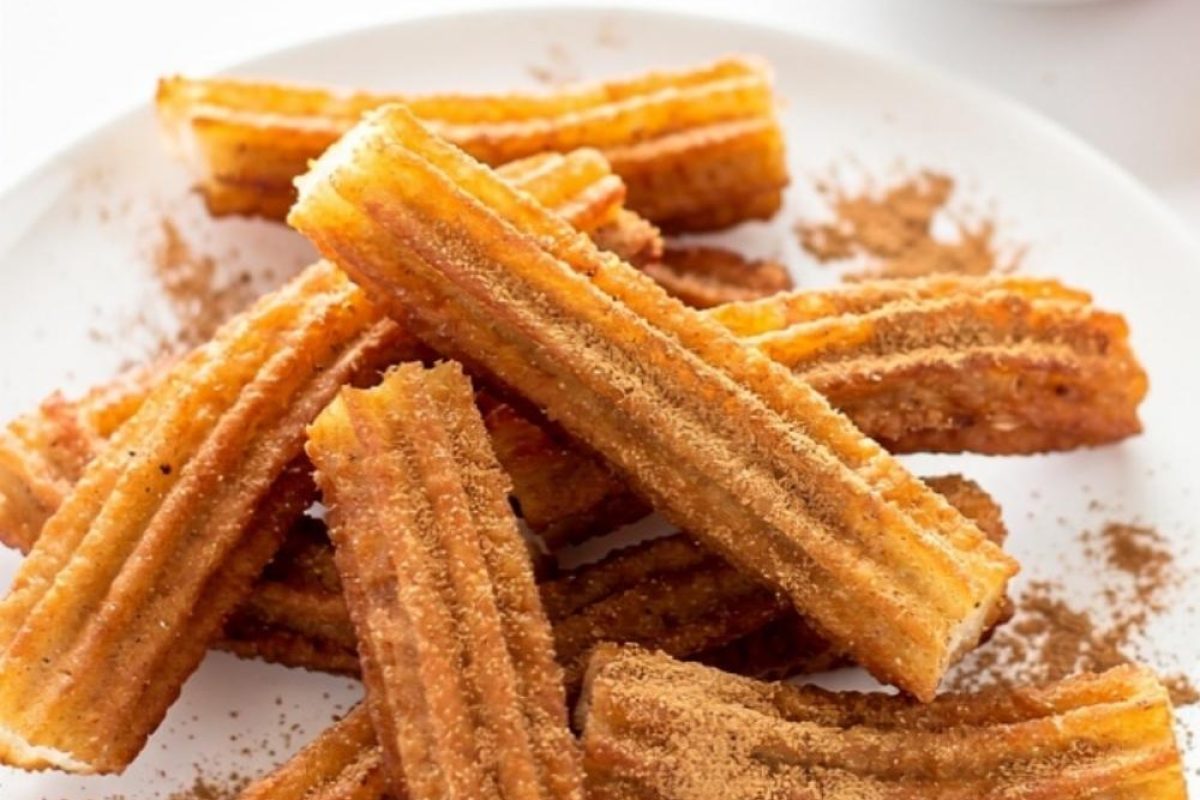 Golden homemade churros arranged on a white ceramic plate dusted with cinnamon sugar, with a small bowl of chocolate dipping sauce on a white table.