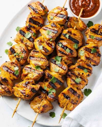 Golden honey chipotle chicken skewers on a white ceramic plate, garnished with fresh cilantro and a small bowl of chipotle sauce on the side, bright natural light on a clean white table.