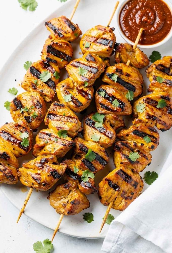 Golden honey chipotle chicken skewers on a white ceramic plate, garnished with fresh cilantro and a small bowl of chipotle sauce on the side, bright natural light on a clean white table.