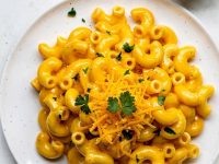 Overhead flat lay of creamy Instant Pot Mac and Cheese on a white ceramic plate with golden cheese sauce, garnished with fresh parsley and shredded cheddar, small bowl of extra cheese sauce on the side, white linen napkin, bright natural daylight on white table