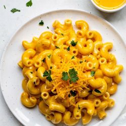 Overhead flat lay of creamy Instant Pot Mac and Cheese on a white ceramic plate with golden cheese sauce, garnished with fresh parsley and shredded cheddar, small bowl of extra cheese sauce on the side, white linen napkin, bright natural daylight on white table