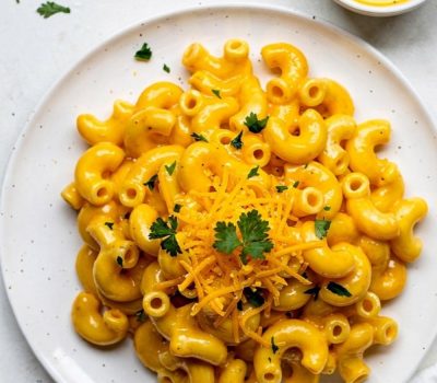 Overhead flat lay of creamy Instant Pot Mac and Cheese on a white ceramic plate with golden cheese sauce, garnished with fresh parsley and shredded cheddar, small bowl of extra cheese sauce on the side, white linen napkin, bright natural daylight on white table