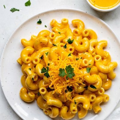 Overhead flat lay of creamy Instant Pot Mac and Cheese on a white ceramic plate with golden cheese sauce, garnished with fresh parsley and shredded cheddar, small bowl of extra cheese sauce on the side, white linen napkin, bright natural daylight on white table