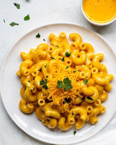 Overhead flat lay of creamy Instant Pot Mac and Cheese on a white ceramic plate with golden cheese sauce, garnished with fresh parsley and shredded cheddar, small bowl of extra cheese sauce on the side, white linen napkin, bright natural daylight on white table