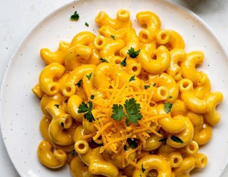 Overhead flat lay of creamy Instant Pot Mac and Cheese on a white ceramic plate with golden cheese sauce, garnished with fresh parsley and shredded cheddar, small bowl of extra cheese sauce on the side, white linen napkin, bright natural daylight on white table