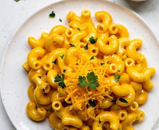 Overhead flat lay of creamy Instant Pot Mac and Cheese on a white ceramic plate with golden cheese sauce, garnished with fresh parsley and shredded cheddar, small bowl of extra cheese sauce on the side, white linen napkin, bright natural daylight on white table