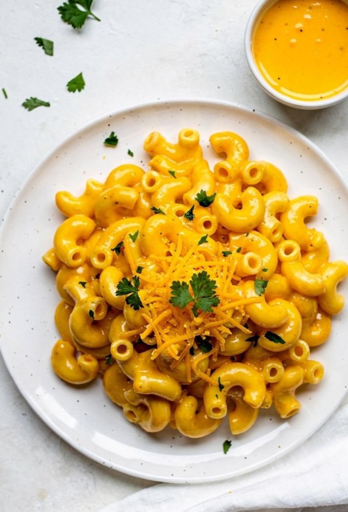 Overhead flat lay of creamy Instant Pot Mac and Cheese on a white ceramic plate with golden cheese sauce, garnished with fresh parsley and shredded cheddar, small bowl of extra cheese sauce on the side, white linen napkin, bright natural daylight on white table