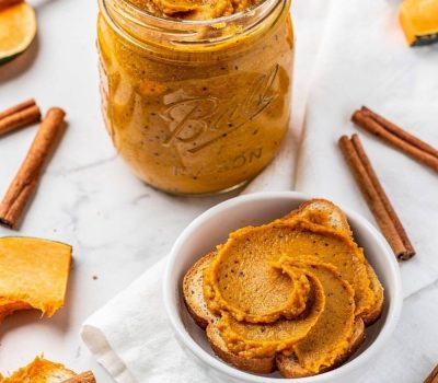 Overhead flat lay of pumpkin butter in a clear glass Mason jar on a white marble table with golden-orange spiced butter visible, spread on toast beside it, cinnamon sticks and fresh pumpkin slices, bright natural light