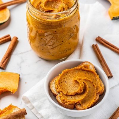 Overhead flat lay of pumpkin butter in a clear glass Mason jar on a white marble table with golden-orange spiced butter visible, spread on toast beside it, cinnamon sticks and fresh pumpkin slices, bright natural light
