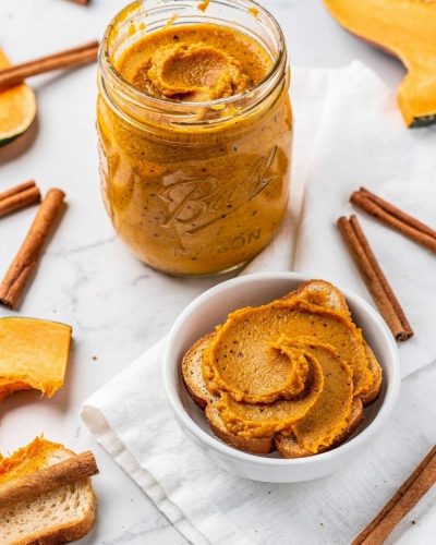 Overhead flat lay of pumpkin butter in a clear glass Mason jar on a white marble table with golden-orange spiced butter visible, spread on toast beside it, cinnamon sticks and fresh pumpkin slices, bright natural light