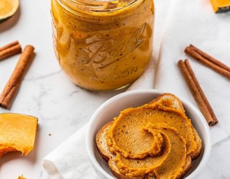 Overhead flat lay of pumpkin butter in a clear glass Mason jar on a white marble table with golden-orange spiced butter visible, spread on toast beside it, cinnamon sticks and fresh pumpkin slices, bright natural light