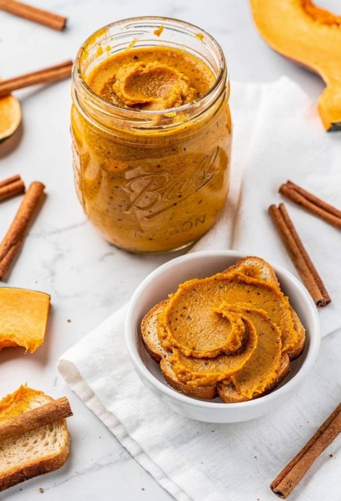 Overhead flat lay of pumpkin butter in a clear glass Mason jar on a white marble table with golden-orange spiced butter visible, spread on toast beside it, cinnamon sticks and fresh pumpkin slices, bright natural light