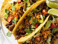 Easy beef tacos recipe served as overhead flat lay on a white ceramic plate with seasoned ground beef, shredded cheese, fresh lettuce, diced tomatoes, and cilantro on a white table
