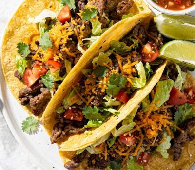 Easy beef tacos recipe served as overhead flat lay on a white ceramic plate with seasoned ground beef, shredded cheese, fresh lettuce, diced tomatoes, and cilantro on a white table