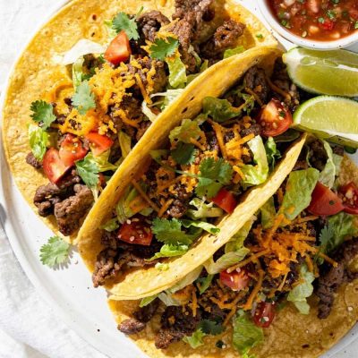 Easy beef tacos recipe served as overhead flat lay on a white ceramic plate with seasoned ground beef, shredded cheese, fresh lettuce, diced tomatoes, and cilantro on a white table