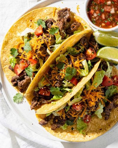 Easy beef tacos recipe served as overhead flat lay on a white ceramic plate with seasoned ground beef, shredded cheese, fresh lettuce, diced tomatoes, and cilantro on a white table