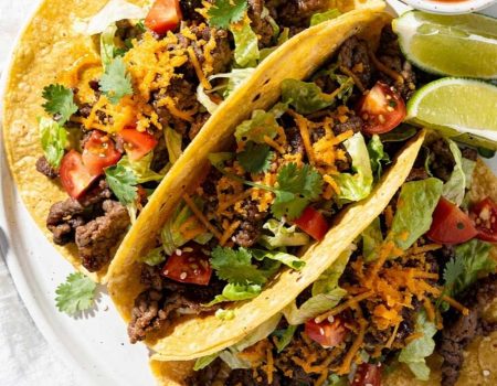 Easy beef tacos recipe served as overhead flat lay on a white ceramic plate with seasoned ground beef, shredded cheese, fresh lettuce, diced tomatoes, and cilantro on a white table