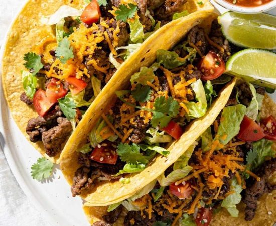Easy beef tacos recipe served as overhead flat lay on a white ceramic plate with seasoned ground beef, shredded cheese, fresh lettuce, diced tomatoes, and cilantro on a white table