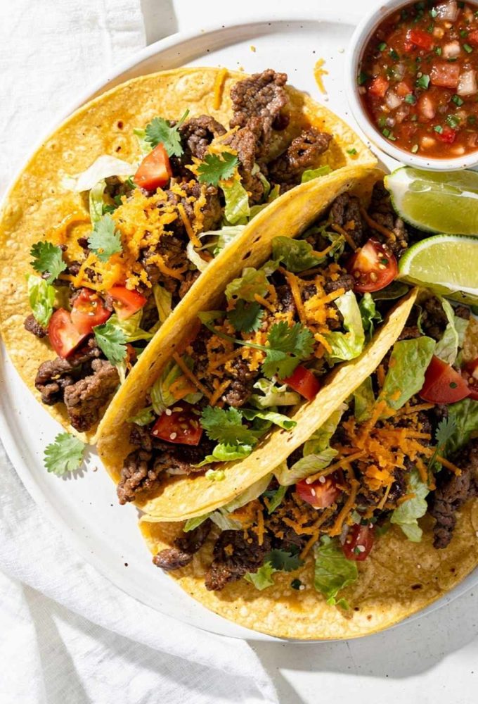Easy beef tacos recipe served as overhead flat lay on a white ceramic plate with seasoned ground beef, shredded cheese, fresh lettuce, diced tomatoes, and cilantro on a white table