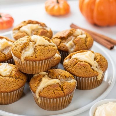 Golden pumpkin cream cheese muffins with cream cheese swirl on a white ceramic plate, white table.