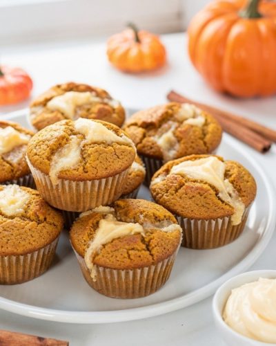 Golden pumpkin cream cheese muffins with cream cheese swirl on a white ceramic plate, white table.