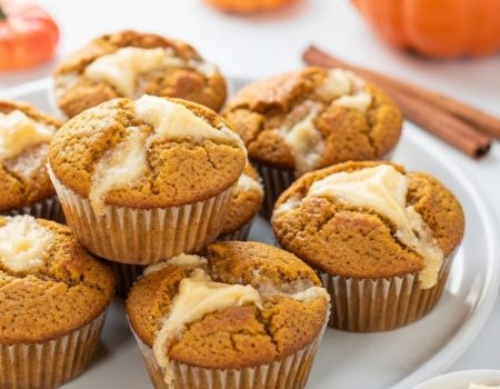 Golden pumpkin cream cheese muffins with cream cheese swirl on a white ceramic plate, white table.