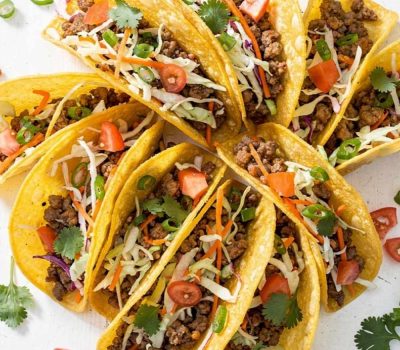 Overhead flat lay of Brandon's Egg Roll Tacos with crispy golden shells, seasoned pork filling, shredded cabbage, carrots, and fresh cilantro on a white table.