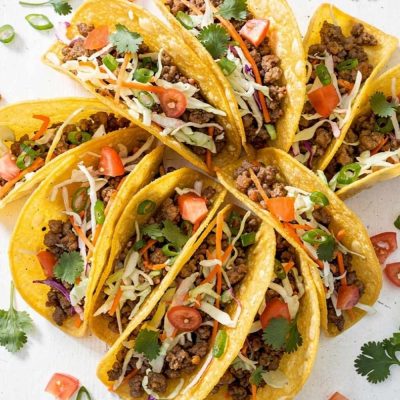 Overhead flat lay of Brandon's Egg Roll Tacos with crispy golden shells, seasoned pork filling, shredded cabbage, carrots, and fresh cilantro on a white table.