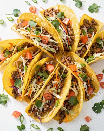 Overhead flat lay of Brandon's Egg Roll Tacos with crispy golden shells, seasoned pork filling, shredded cabbage, carrots, and fresh cilantro on a white table.