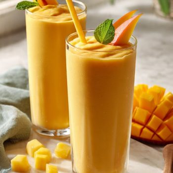 Two tall glasses of tropical fruit smoothie with milk garnished with fresh mango and mint on white marble — best 5-minute blender recipe