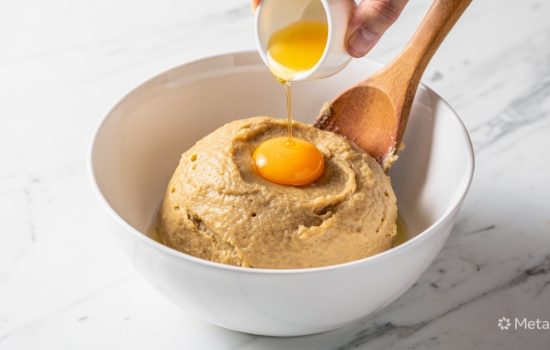 Smooth homemade churro dough in a white ceramic mixing bowl with an egg being added on a white marble surface.