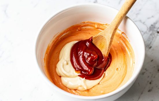 Step 2 — mixing mayonnaise and ketchup together in a white bowl with a wooden spoon on a white marble surface, horizontal food photography.